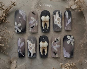 May include: A set of ten press-on nails featuring a dark gray and beige gradient base. The designs include 3D white flower petals, black ants, realistic 3D human teeth, and white bone-like structures. The text Art Therapy is printed at the top.