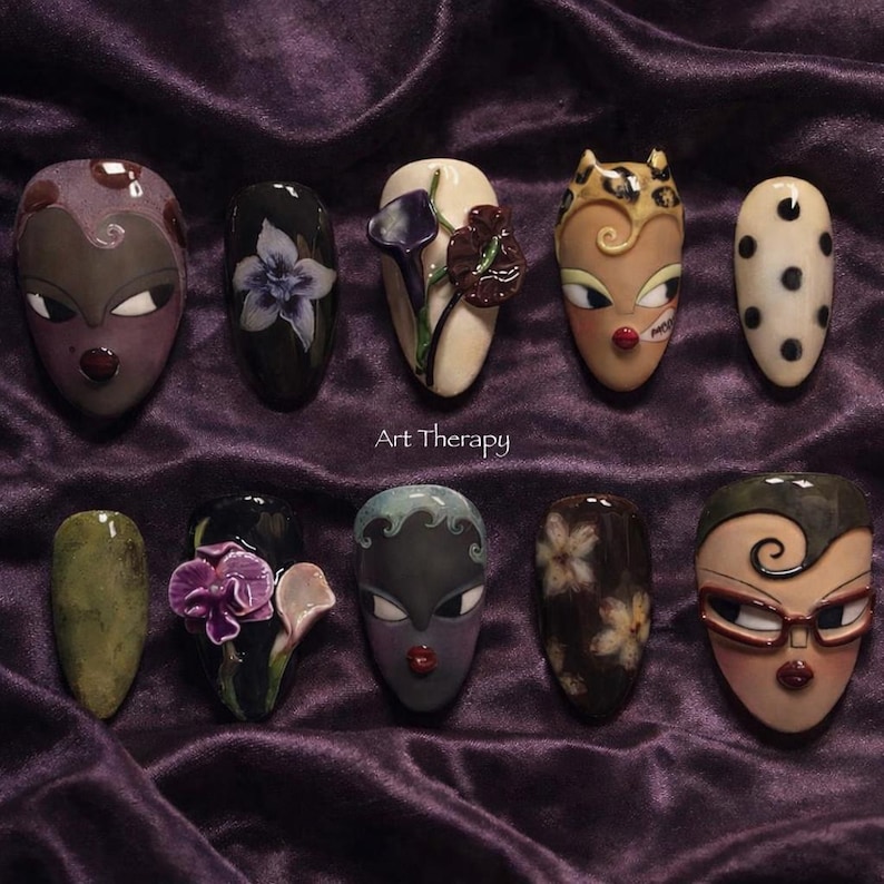 May include: A collection of ten uniquely designed nail art pieces. Each nail features a different design, including floral patterns, stylized faces, and abstract art. The nails are displayed on a purple velvet surface, with the text "Art Therapy" visible.