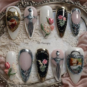 May include: A collection of ten long, oval-shaped artificial nails. The nails feature various designs, including floral accents, sword-like embellishments, and framed miniature paintings. The text "Art Therapy" is visible on the surface.