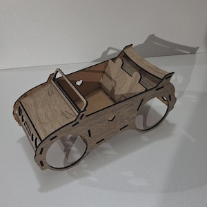 Puede incluir: Un coche de juguete de madera con diseño descapotable. El coche es de madera marrón claro con contornos negros y presenta detalles recortados, incluyendo una forma de corazón y huecos para las ruedas. El interior tiene dos asientos y un marco de parabrisas.