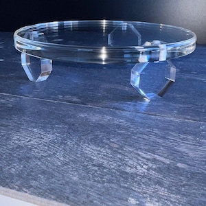 May include: A clear acrylic display stand featuring a circular top platform supported by three distinct hexagonal-shaped legs. The transparent material provides a sleek, neutral look, while the geometric base design offers stability for the elevated surface.