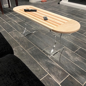 May include: A modern coffee table with a light wood top featuring a striped design and rounded edges. The table is supported by clear, curved acrylic legs. A remote control and a small black device rest on the table's surface.