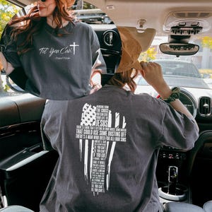 May include: Dark gray t-shirt with white text and a cross graphic. The front of the shirt reads "Til' You Can't" and the back features a cross design with the text "Hanging From The Cross You Can Give Your Life To Jesus".