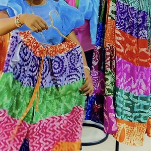 May include: A vibrant patchwork skirt with a drawstring waist is displayed on a gold hanger. The skirt showcases panels of purple, green, and pink fabric with intricate patterns. Other skirts with similar designs are visible in the background.