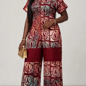 May include: A short-sleeved jumpsuit featuring a deep burgundy base colour with a repeating geometric pattern in shades of light blue, cream, and dark red. The garment has a round neckline, a straight-leg silhouette, and a solid burgundy panel.