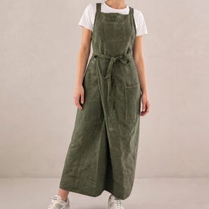 Puede incluir: Mono de lino verde oliva con cuello cuadrado, perneras anchas y cinturón de tela a juego. El mono tiene un bolsillo delantero y un corte holgado. La modelo lleva una camiseta blanca debajo y zapatillas blancas.