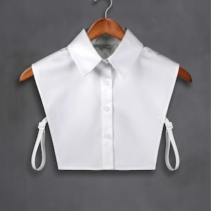 Puede incluir: Un cuello de camisa blanco desmontable con un diseño clásico abotonado. El cuello está hecho de una tela lisa y presenta un cuello puntiagudo. Las correas ajustables son visibles a cada lado, y el cuello cuelga de una percha de madera.