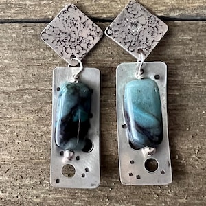May include: A pair of handcrafted earrings featuring a square, textured silver top and a rectangular silver base. Each earring has a vibrant, blue-green stone with dark swirls. The earrings are suspended from a silver hook.