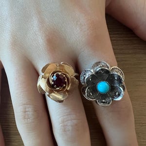 May include: Two flower-shaped rings. One is gold-toned with a dark red center stone. The other is silver-toned with a turquoise blue center stone. Both rings are on a hand.