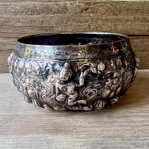 May include: A silver-colored bowl with intricate relief carvings of figures and foliage. The bowl has a wide, rounded shape with a detailed rim. The carvings depict human figures in various poses, creating a detailed and ornate design.
