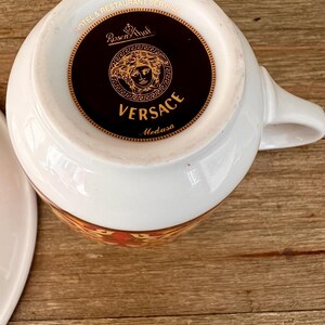 Puede incluir: Taza de t&eacute; blanca con asa, con un dise&ntilde;o floral dorado y naranja alrededor del borde. La base de la taza muestra el logotipo de Versace Medusa en dorado sobre un fondo negro, con las palabras "VERSACE" y "Medusa".