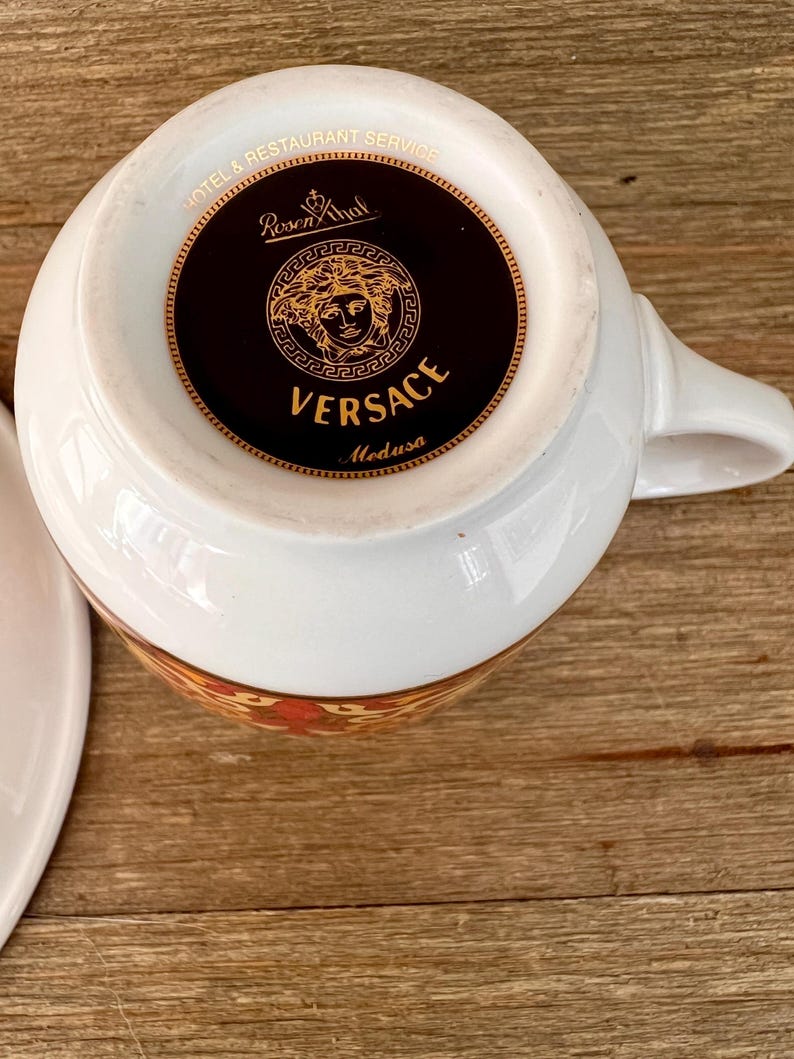 Puede incluir: Taza de t&eacute; blanca con asa, con un dise&ntilde;o floral dorado y naranja alrededor del borde. La base de la taza muestra el logotipo de Versace Medusa en dorado sobre un fondo negro, con las palabras "VERSACE" y "Medusa".