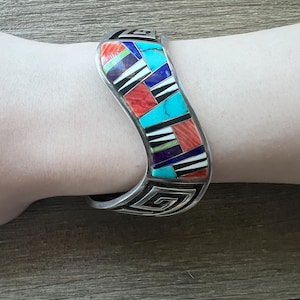 May include: A silver cuff bracelet with a mosaic design. The mosaic features red, turquoise, black, white, purple, and blue geometric shapes. The bracelet has a curved shape and a decorative pattern on the edges.