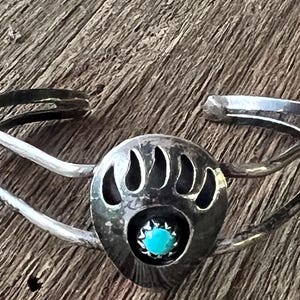 May include: A silver cuff bracelet featuring a central teardrop-shaped plaque with a cutout bear paw print design. A small, round, bright turquoise stone is set in the center of the paw print. The bracelet has a double-band open design.
