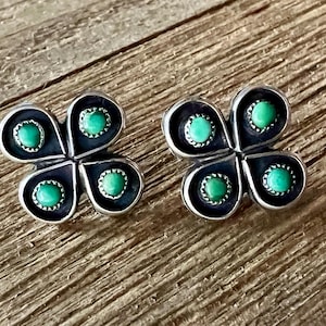 May include: A pair of stud earrings featuring a four-petal flower design crafted from dark oxidized metal. Each petal contains a small, round, bright green turquoise stone set within a serrated bezel, creating a distinct contrast against the dark metal finish.
