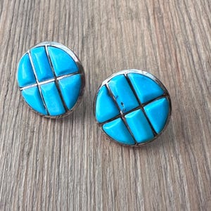 May include: A pair of round turquoise stud earrings. Each earring features a bright blue, geometric design set in a silver-colored metal. The earrings are displayed on a light brown wooden surface.