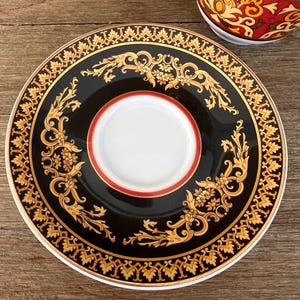 Puede incluir: Un platillo decorativo negro y dorado con un centro blanco y un borde rojo. El platillo presenta un dise&ntilde;o floral dorado ornamentado alrededor del borde y el centro. Una porci&oacute;n de una taza de t&eacute; a juego es visible en la esquina superior derecha.