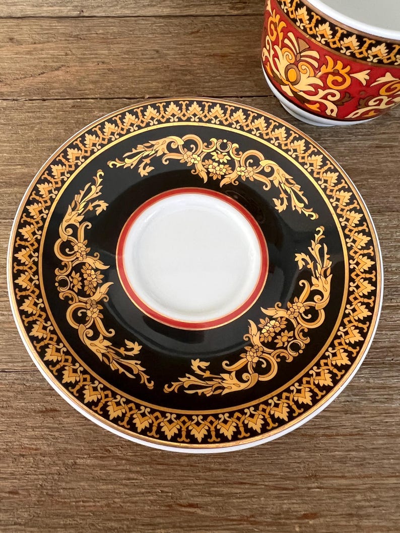 Puede incluir: Un platillo decorativo negro y dorado con un centro blanco y un borde rojo. El platillo presenta un dise&ntilde;o floral dorado ornamentado alrededor del borde y el centro. Una porci&oacute;n de una taza de t&eacute; a juego es visible en la esquina superior derecha.