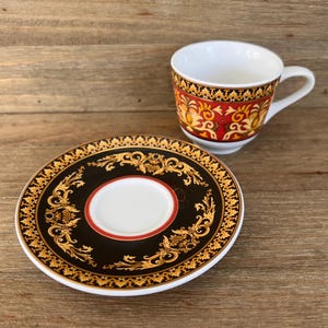 Puede incluir: Un juego de taza y platillo decorativo. La taza tiene un interior y un asa blancos, con una banda estampada en rojo y dorado. El platillo es negro con un dise&ntilde;o floral dorado y un centro blanco.