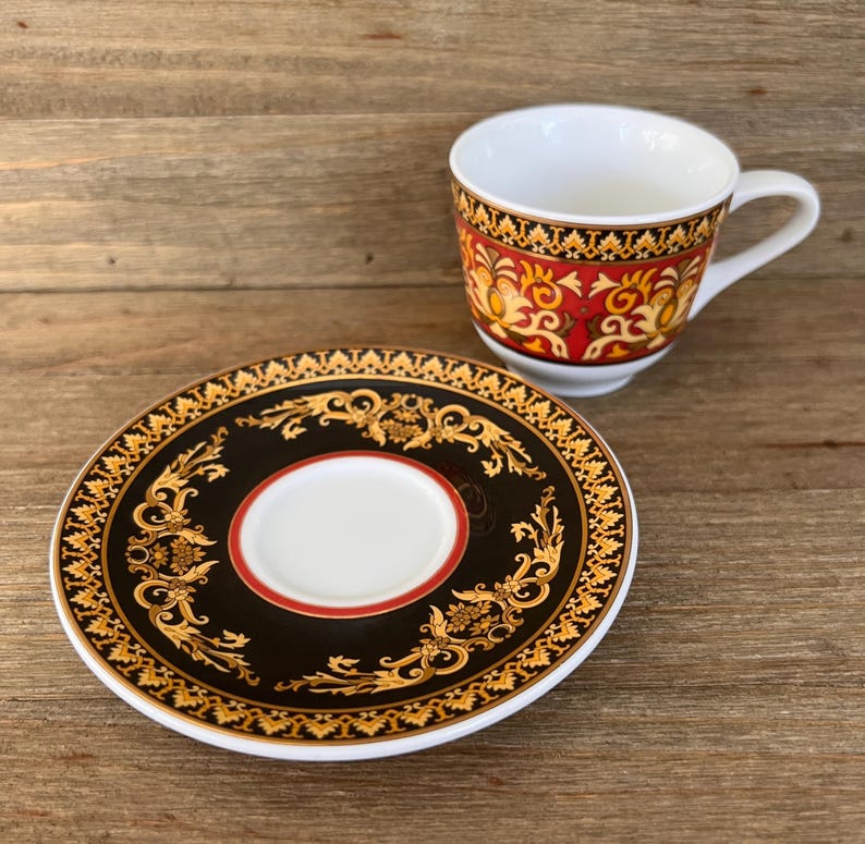 Puede incluir: Un juego de taza y platillo decorativo. La taza tiene un interior y un asa blancos, con una banda estampada en rojo y dorado. El platillo es negro con un dise&ntilde;o floral dorado y un centro blanco.