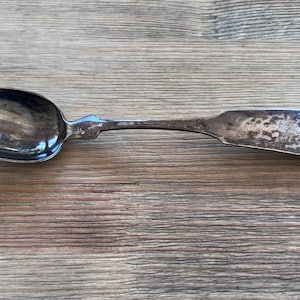 May include: A vintage silver spoon with a tarnished finish, resting on a wooden surface. The spoon features a rounded bowl and a detailed handle. The spoon's design suggests it is an antique or a piece of historical silverware.