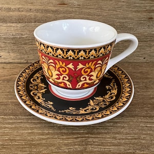 May include: A white teacup and saucer set with a decorative design. The cup and saucer feature a black, red, and gold pattern. The cup has a white handle and interior. The saucer is round with a matching design.