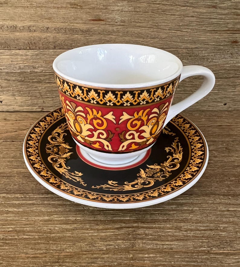 Puede incluir: Juego de taza y platillo decorativo. La taza y el platillo presentan un dise&ntilde;o negro, rojo y dorado. La taza tiene un asa e interior blancos. El platillo es redondo con un dise&ntilde;o a juego. Hecho de cer&aacute;mica.