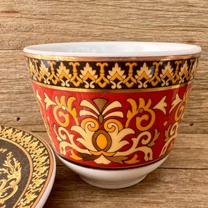 Puede incluir: Una taza de cer&aacute;mica decorativa con un platillo a juego. La taza presenta un fondo rojo con patrones florales dorados y amarillos. El borde est&aacute; acentuado con una banda dorada y un dise&ntilde;o negro y amarillo.