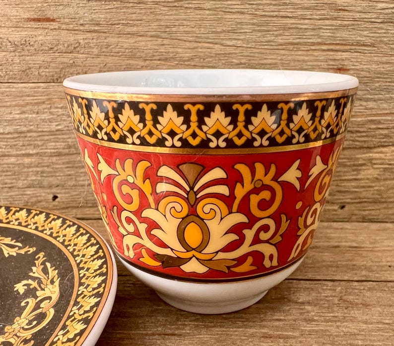 Puede incluir: Una taza de cer&aacute;mica decorativa con un platillo a juego. La taza presenta un fondo rojo con patrones florales dorados y amarillos. El borde est&aacute; acentuado con una banda dorada y un dise&ntilde;o negro y amarillo.