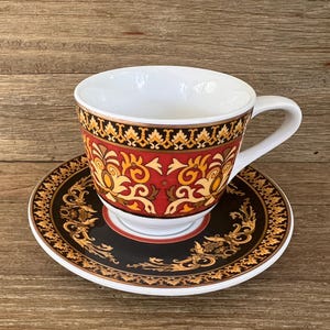 Puede incluir: Una taza de t&eacute; blanca con asa y platillo a juego. La taza y el platillo presentan un patr&oacute;n decorativo en rojo, negro y dorado. La taza de t&eacute; est&aacute; sobre una superficie de madera.