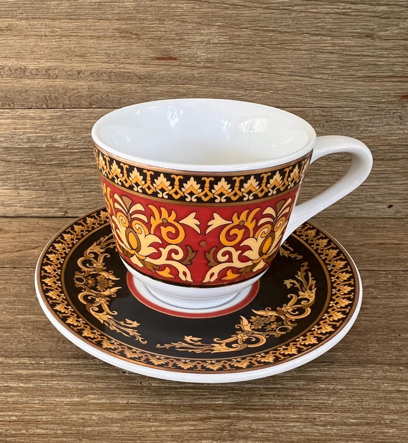 Puede incluir: Una taza de t&eacute; blanca con asa y platillo a juego. La taza y el platillo presentan un patr&oacute;n decorativo en rojo, negro y dorado. La taza de t&eacute; est&aacute; sobre una superficie de madera.