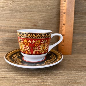 Puede incluir: Un juego de taza y platillo decorativo con un patr&oacute;n rojo y dorado. La taza tiene un asa blanca y se apoya en un platillo a juego. Una regla de madera est&aacute; al lado de la taza. El juego est&aacute; sobre una superficie de madera. La taza mide unos 6 cm de altura.