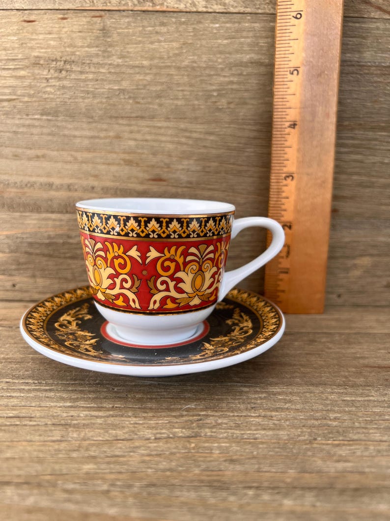Puede incluir: Un juego de taza y platillo decorativo con un patr&oacute;n rojo y dorado. La taza tiene un asa blanca y se apoya en un platillo a juego. Una regla de madera est&aacute; al lado de la taza. El juego est&aacute; sobre una superficie de madera. La taza mide unos 6 cm de altura.