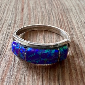 May include: A silver ring with a wide band featuring a mosaic of blue and green iridescent stones. The ring is set against a wooden surface, showcasing its vibrant colors and unique design. A piece of jewelry.