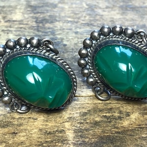 May include: A pair of green carved stone earrings with silver settings. The earrings have a beaded border and a dangling hook.