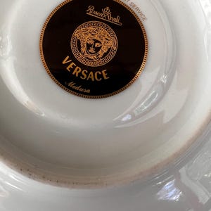 Puede incluir: Plato de porcelana blanca con un logotipo Versace Medusa dorado y negro. El logotipo presenta el nombre Versace y la marca Rosenthal. El plato est&aacute; dise&ntilde;ado para el servicio de hosteler&iacute;a y restauraci&oacute;n.