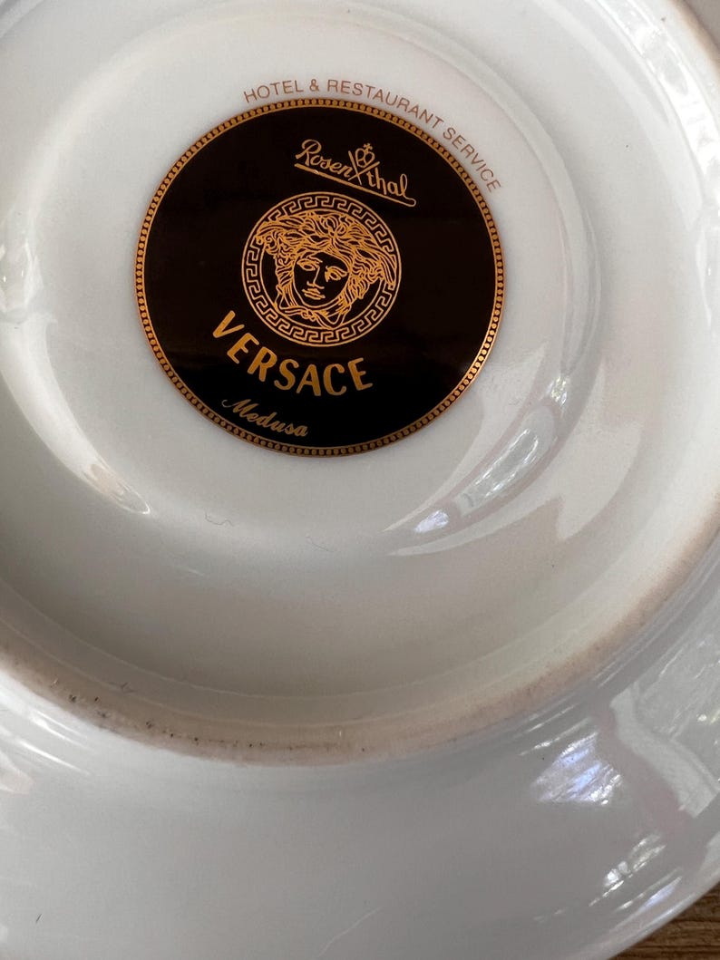 Puede incluir: Plato de porcelana blanca con un logotipo Versace Medusa dorado y negro. El logotipo presenta el nombre Versace y la marca Rosenthal. El plato est&aacute; dise&ntilde;ado para el servicio de hosteler&iacute;a y restauraci&oacute;n.