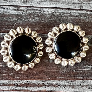 May include: A pair of silver-toned stud earrings with a large, round, glossy black center stone. The earrings have a decorative border of small, ridged silver beads. The earrings are displayed on a weathered wooden surface.