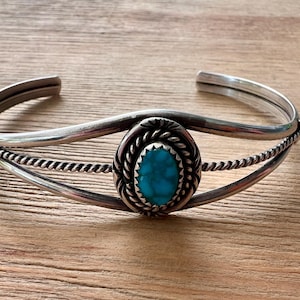 May include: A silver cuff bracelet with a central oval turquoise stone. The turquoise is surrounded by a twisted rope design, and the bracelet has a simple, elegant design. The bracelet is displayed on a wooden surface.