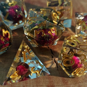 May include: A collection of handmade resin dice, each containing dried flowers and gold flakes. The dice are transparent with gold numerals and a red flower. The set includes various shapes, such as a pyramid and a 20-sided die.