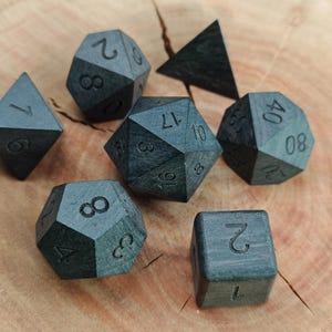 Könnte beinhalten: Eine Sammlung dunkelgrauer Würfel aus Holz für Tabletop-Spiele. Das Set enthält verschiedene Formen, wie einen Würfel, ein Tetraeder und einen 20-seitigen Würfel, jeweils mit eingravierten Zahlen. Die Würfel liegen auf einer hellbraunen Holzoberfläche.