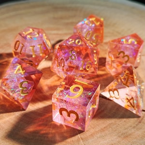 May include: A set of seven resin dice in pink and orange hues, with gold numerals. The transparent dice contain iridescent glitter. The dice are displayed on a wooden surface, with shadows from the light.