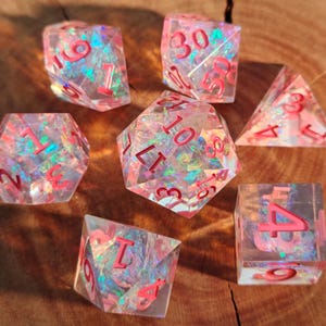May include: A set of seven clear resin dice with iridescent glitter and red numbering. The dice include a D4, D6, D8, D10, D12, D20, and a percentile die. The dice are designed for tabletop role-playing games.