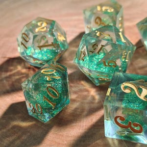 May include: A collection of polyhedral dice with a clear, teal-colored resin. The dice have gold-colored numbers and are filled with green glitter. The dice are designed for tabletop gaming.