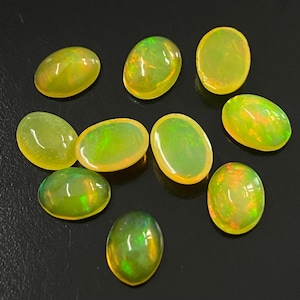 Può includere: Una collezione di nove opali cabochon di forma ovale, ognuno dei quali presenta un colore di base giallo-verde traslucido con lampi interni di fuoco arancione, verde e giallo. Le pietre preziose sono lisce, levigate e disposte in modo sparso.