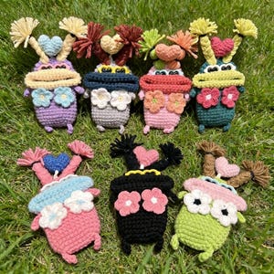 May include: A collection of handmade crochet dolls in various colors, including purple, gray, pink, and green. Each doll features a unique design with heart-shaped accents, flower details, and textured yarn hair. The dolls are arranged on a bed of green grass.