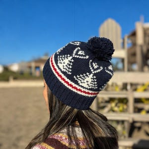 May include: Navy blue knit beanie with a white anchor pattern, a red and white striped band, and a dark blue pom-pom. The hat is worn by a person with long brown hair.