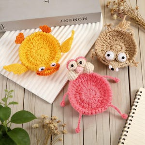 Puede incluir: Tres posavasos de ganchillo hechos a mano con forma de criaturas caprichosas. Los posavasos incluyen un pato amarillo con detalles en naranja, una criatura rosa con patas largas y una criatura marrón con ojos grandes.