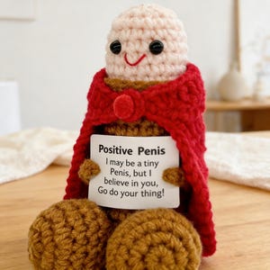 May include: A crocheted doll in the shape of a penis, wearing a red cape. The doll has a smiling face and holds a sign that reads "Positive Penis". The doll is made of beige and brown yarn.