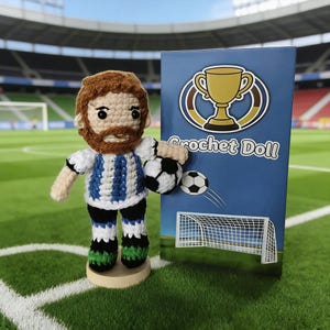 May include: A handmade crochet doll of a soccer player, holding a soccer ball. The doll wears a white and blue striped jersey, black shorts, and green and black shoes. A blue box with a trophy and the words "Crochet Doll" is behind the doll.